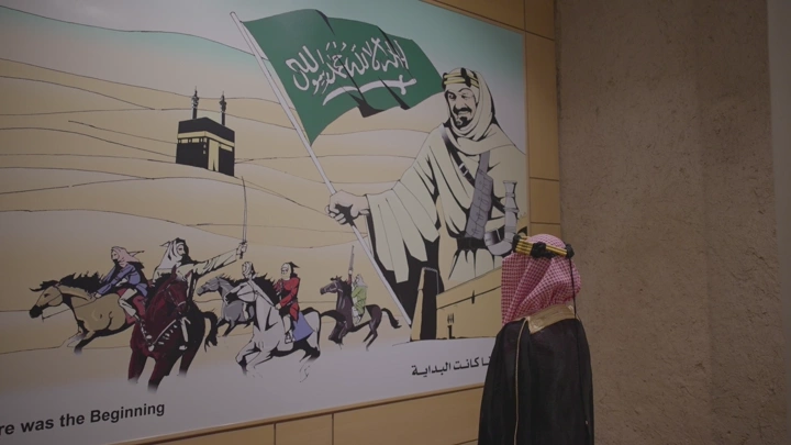 لوحة تاريخية وشخص واقف امامه بالزي السعودي ليوم التاسيس في قصر المصمك بالرياض، تصوير داخلي.