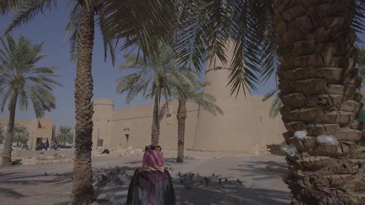 قصر المصمك في الرياض محاط بأشجار النخيل، شخص يمشي ولابس بشت وعقال يوم التاسيس لقطة نهارية.