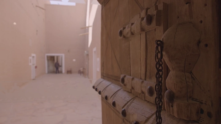 باب خشبي تقليدي مع شخص يمشي ويرتدي الزي السعودي ليوم التاسيس في قصر المصمك بالرياض، لقطة داخلية.