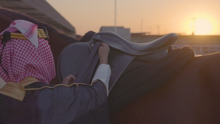 شاب يشد السرج على الخيل عند الغروب ويرتدي الزي السعودي ليوم التاسيس، لقطة قريبة.