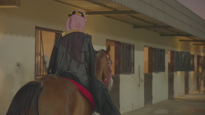 شاب بلباس سعودي يمتطي فرس عربي أمام إسطبل، لقطة خلفية.