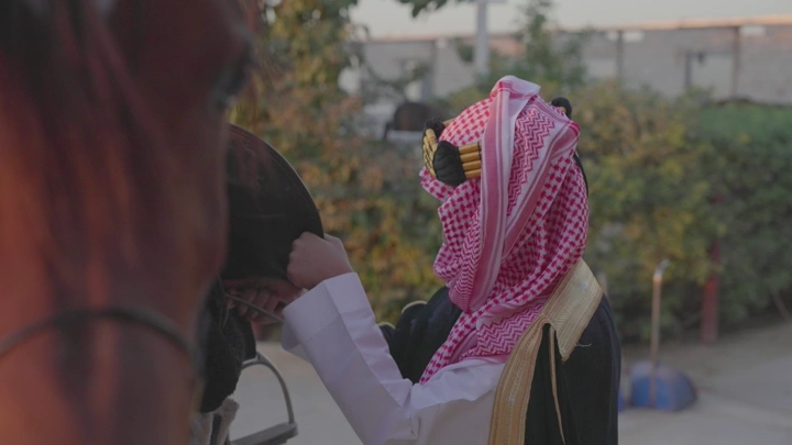 شاب يرتدي الزي التقليدي ليوم التاسيس يشد السرج على الخيل في الخارج، لقطة قريبة.