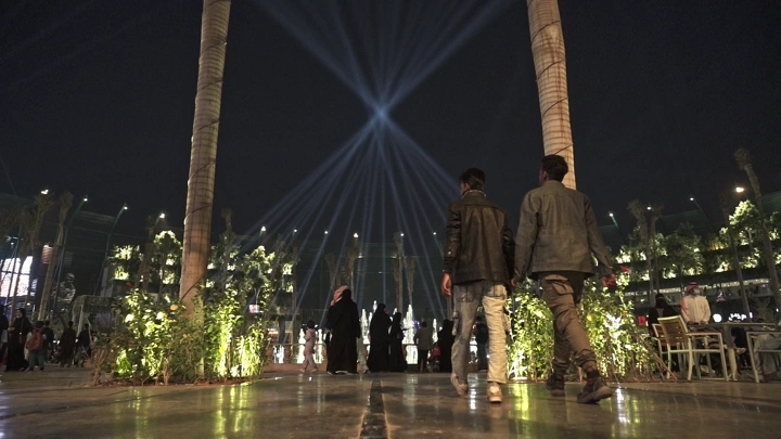 أشخاص يمشون في البوليفارد الرياض ليلاً، إضاءة مبهرة.