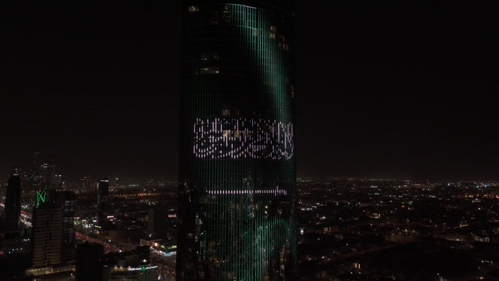 برج في الرياض مضاء بعلم السعودية ليلاً، لقطة جوية.