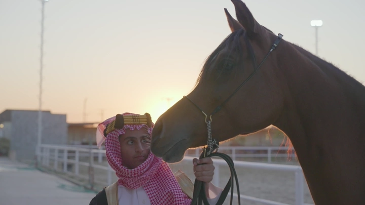 شاب يرتدي الزي السعودي يقف بجانب خيل عند الغروب، لقطة قريبة.