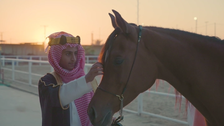 شاب يرتدي الزي السعودي يقف بجانب خيل عند الغروب، لقطة قريبة.