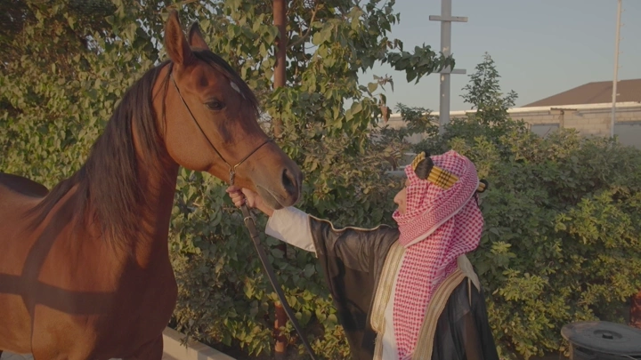 شاب يرتدي الزي السعودي يقف بجانب خيل في حديقة، لقطة نهارية.