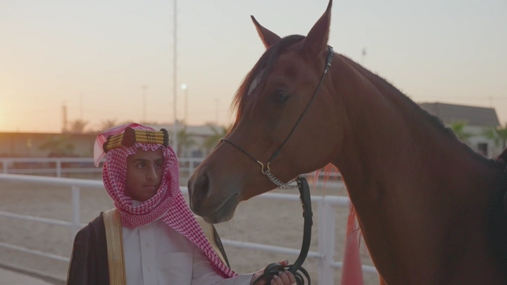 شاب يرتدي الزي السعودي يقف بجانب خيل في مضمار عند الغروب.