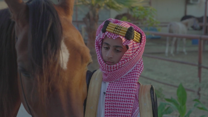 شاب يرتدي الزي السعودي بجانب خيل في مضمار، لقطة مقربة.