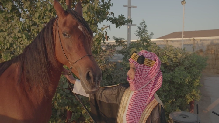 شاب يرتدي الزي السعودي يقف بجانب خيل في مزرعة، لقطة نهارية.