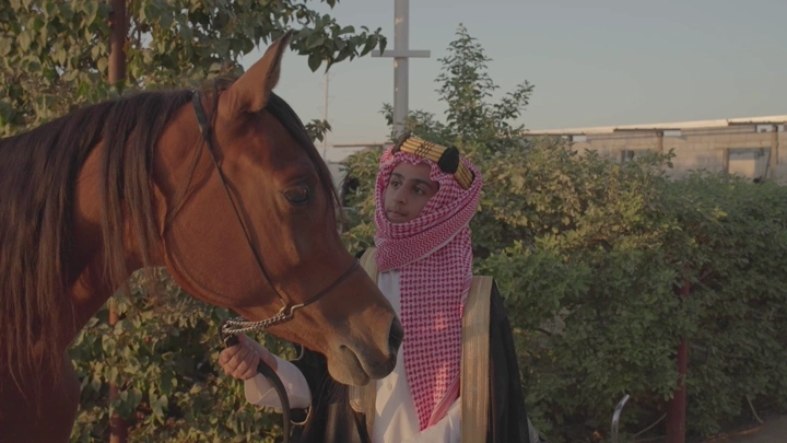 شاب يرتدي الزي التقليدي السعودي ليوم التاسيس ينظر إلى حصان في مزرعة، لقطة نهارية.
