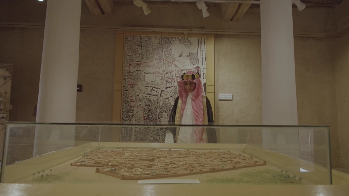 شخص يرتدي الزي السعودي ينظر إلى نموذج مصغر في قصر المصمك، تصوير داخلي.