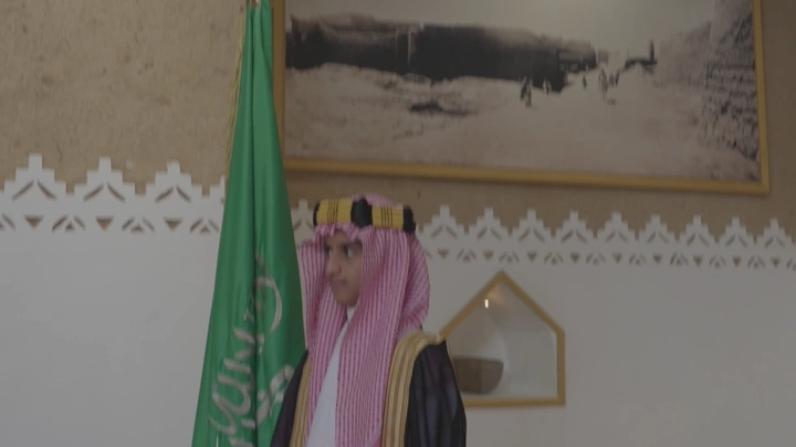 رجل يرتدي الزي السعودي بجانب علم السعودية في قصر المصمك، لقطة داخلية.