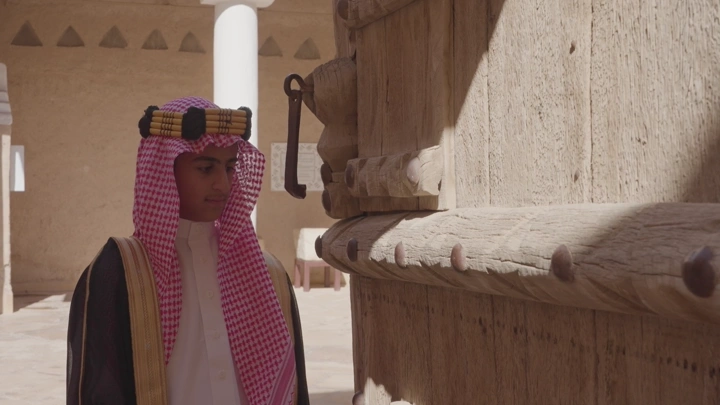 شاب يرتدي الزي السعودي يقف أمام باب قصر المصمك في الرياض، لقطة نهارية.