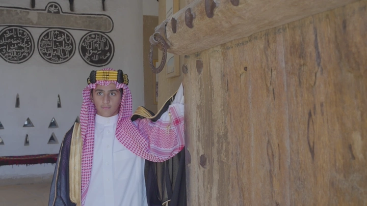 شاب يرتدي الزي السعودي بجانب باب خشبي في قصر المصمك، لقطة داخلية.