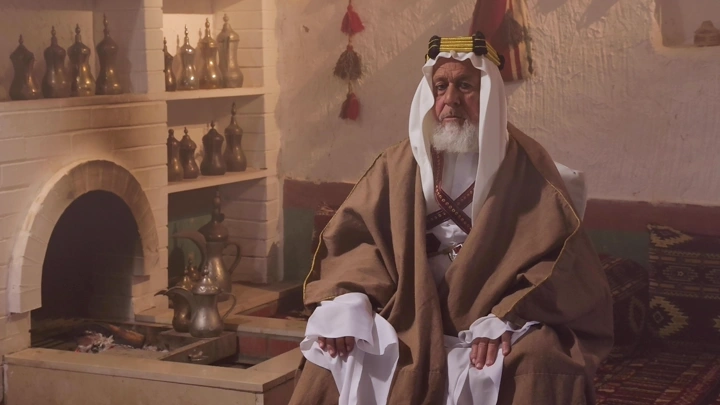 رجل مسن بلباس تراثي سعودي في غرفة شعبية، لقطة داخلية.