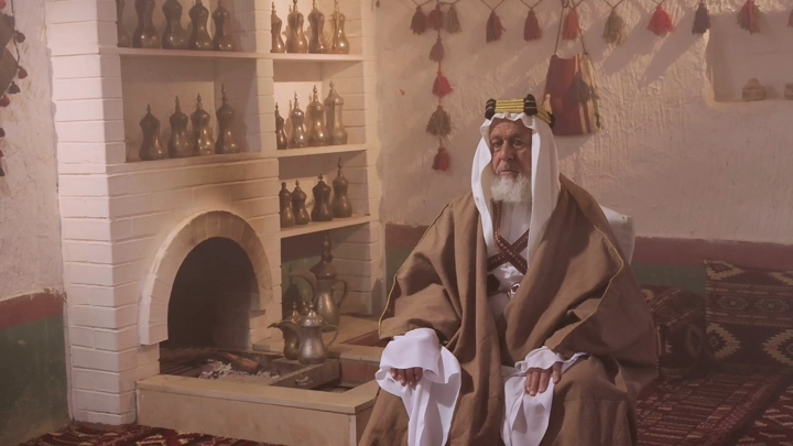 رجل مسن بلباس تراثي سعودي في غرفة تقليدية، لقطة داخلية.
