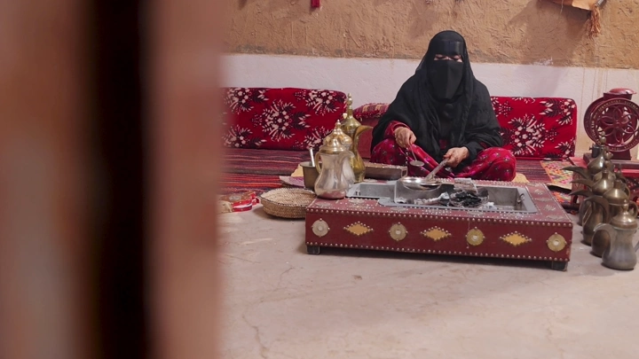 امرأة بلباس تراثي سعودي تحمس القهوة في مجلس عربي، لقطة داخلية.