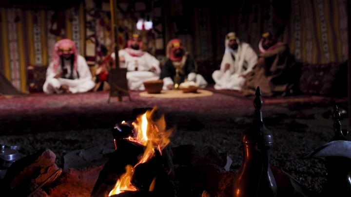 رجال بلباس تراثي سعودي في خيمة بجانب الحطب، لقطة ليلية.