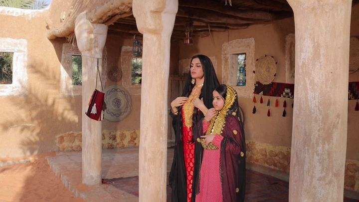 فتاة وأمها بلباس تراثي سعودي في بيت طيني، لقطة نهارية.