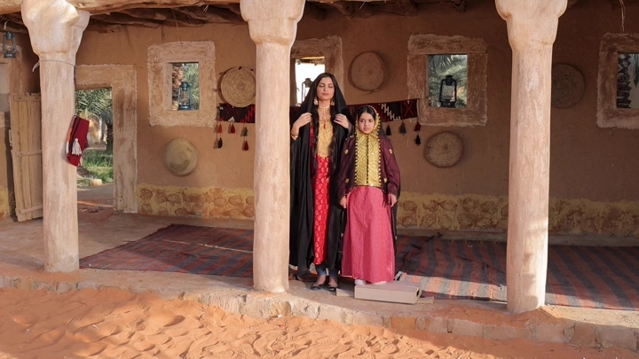 فتاة وأمها بلباس تراثي سعودي في بيت طيني، لقطة نهارية.