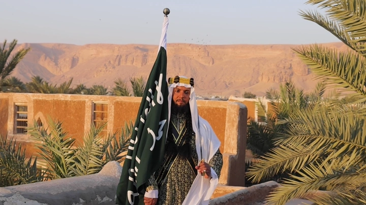رجل بلباس سعودي يحمل علم السعودية القديم في واحة صحراوية، لقطة نهارية.