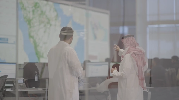 رجال يتحدثون في مكان عمل وامامهم خريطة المملكة العربية السعودية، لقطة من الخلف.