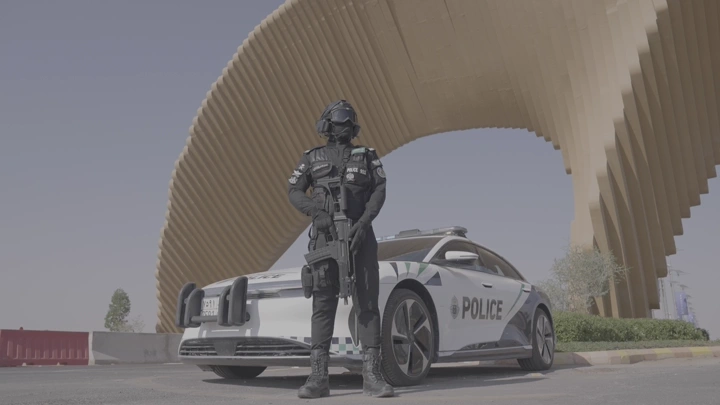 عسكري سعودي يقف بجانب سيارة شرطة عند بوابة الصقور في ملهم في الرياض، لقطة نهارية.