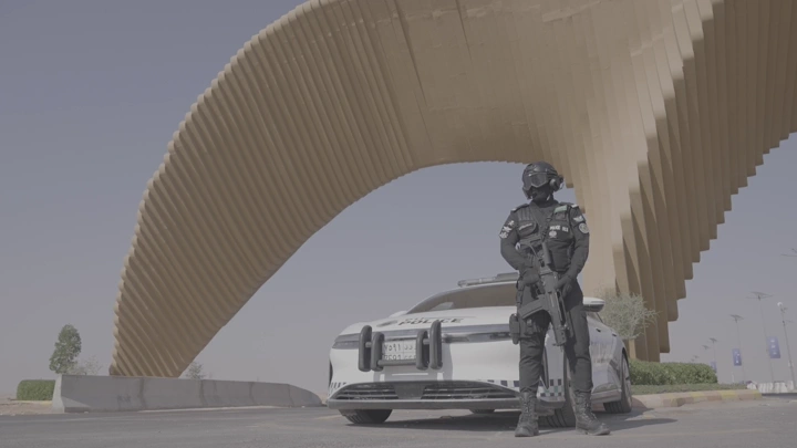 عسكري سعودي يقف بجانب سيارة شرطة عند بوابة الصقور في ملهم في الرياض، لقطة نهارية.