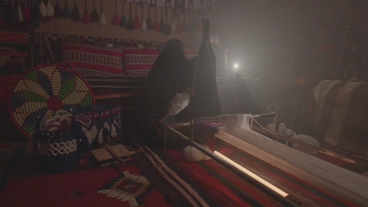 امرأة تنسج السدو في غرفة تقليدية، إضاءة خافتة.