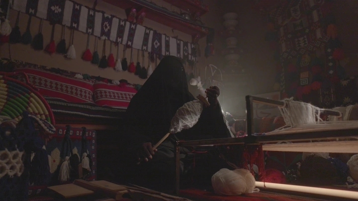 امرأة تنسج السدو في غرفة تقليدية، إضاءة خافتة.