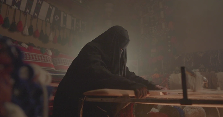 امرأة تنسج على النول في غرفة تقليدية، إضاءة خافتة.