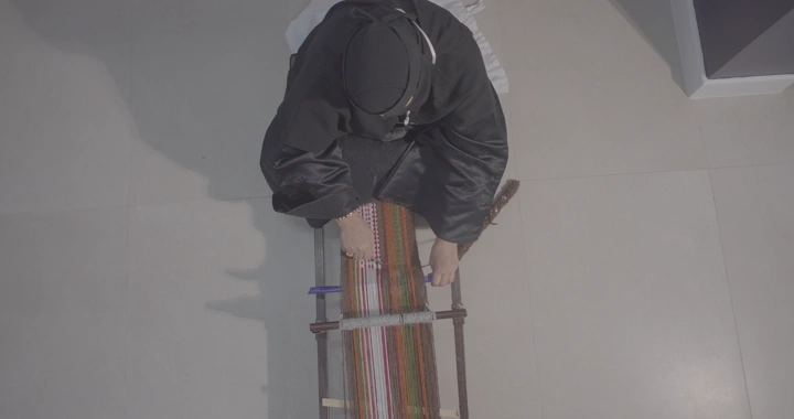 حرفية تعمل على نول السدو في غرفة مضاءة، لقطة من الأعلى.