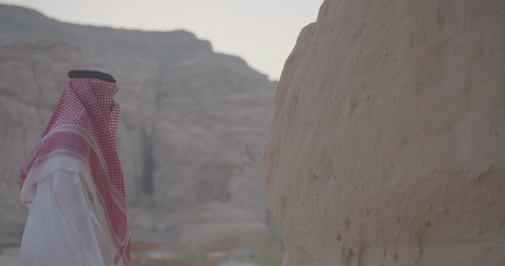رجل يرتدي الزي التقليدي يقف أمام جبال العلا ويلمس الجبال، لقطة جانبية.