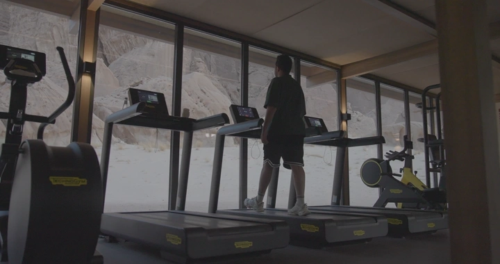 رجل على جهاز مشي في صالة رياضية تطل على جبال العلا، لقطة داخلية.