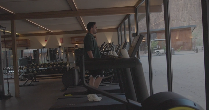 شخص على جهاز سير في صالة رياضية تطل على جبال العلا، لقطة داخلية.