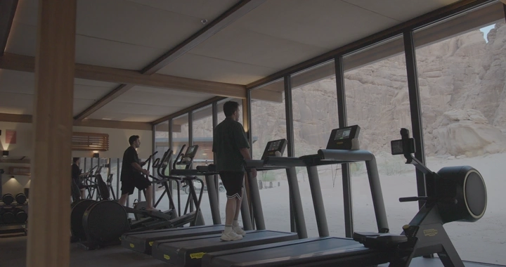شخص على جهاز سير في صالة رياضية تطل على جبال العلا، لقطة داخلية.