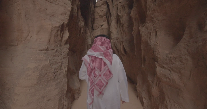 شخص يرتدي ثوبًا وغترة يمشي بين صخور العلا، لقطة خلفية.