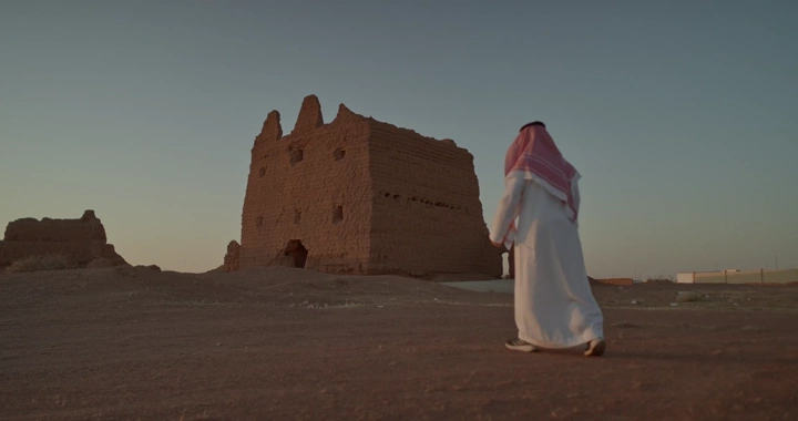 رجل سعودي أمام بيت طيني في الصحراء، لقطة من الخلف.