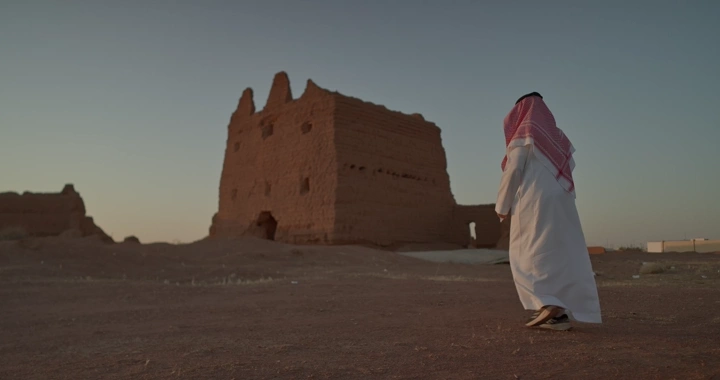 رجل سعودي أمام بيت طيني قديم في الصحراء، لقطة من الخلف.