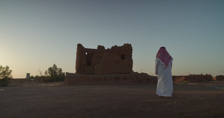 رجل سعودي أمام بيت طين في الصحراء، لقطة من الخلف.