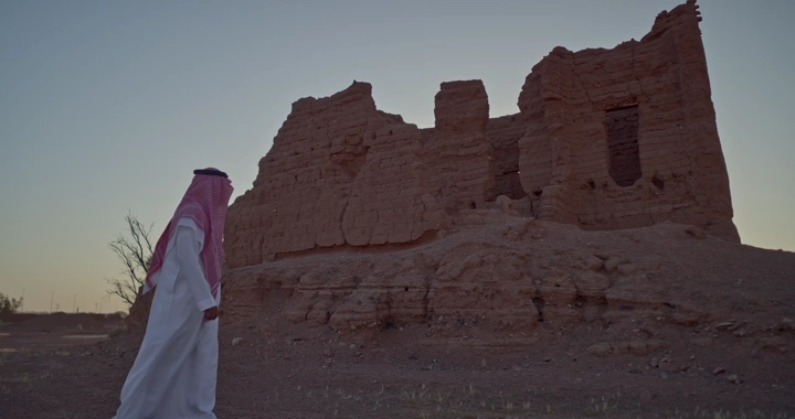 رجل سعودي أمام بيت طيني قديم عند الغروب، لقطة واسعة.