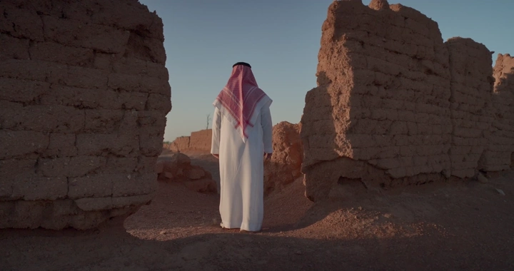 رجل يرتدي ثوبًا وغترة يقف بين جدران طينية، لقطة خلفية.