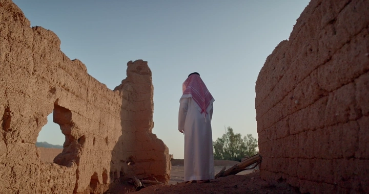 رجل يقف أمام بيت طيني قديم، لقطة من الخلف.