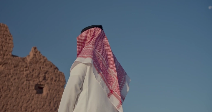 رجل يرتدي شماغ يقف أمام بيت طيني، تصوير نهاري.