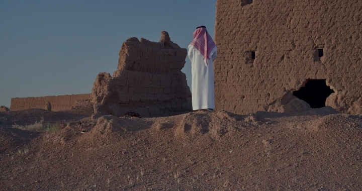 رجل يرتدي ثوبًا يقف أمام بيت طيني قديم، لقطة من الخلف.