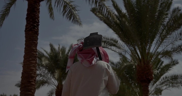 مزارع يرتدي شماغًا في بستان نخيل ويحمل المجرفه، لقطة من الخلف.