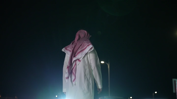 شخص يرتدي ثوبًا يمشي على طريق المشاة امام سيارات، لقطة من الخلف.