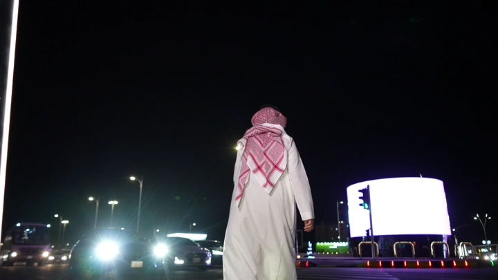 رجل يرتدي ثوباً وغترة يسير على ممر مشاة امام السيارات ليلاً، لقطة من الخلف.