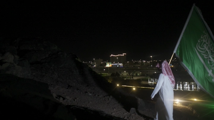 شخص يحمل العلم السعودي في الليل، منظر المدينة في الخلفية.